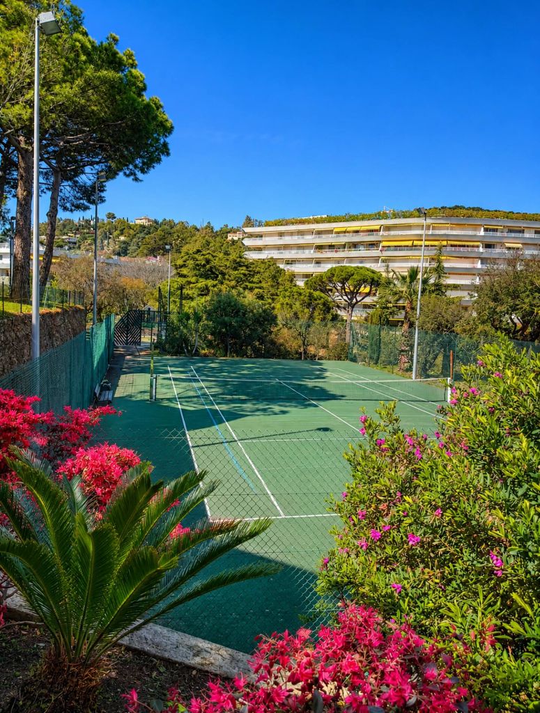 Court de tennis en résine verte de la résidence Villa Francia, entouré de pins, de lauriers-roses et de massifs fleuris