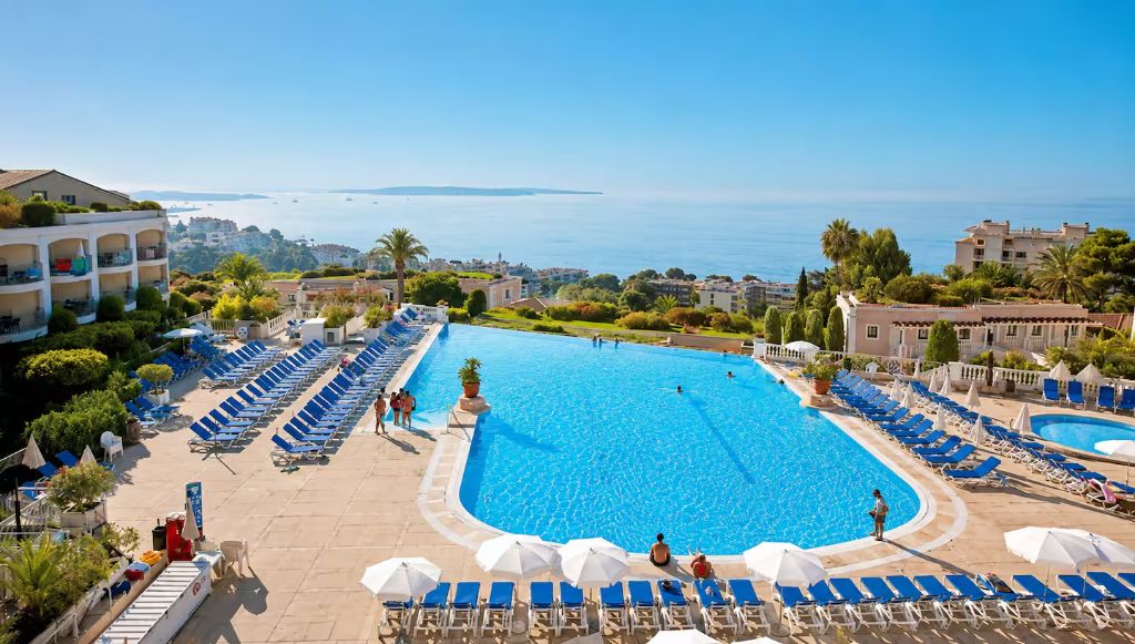 Vue d'ensemble de la piscine de la résidence Villa Francia à Cannes, entourée de transats bleus, avec la mer et les îles de Lérins à l'horizon